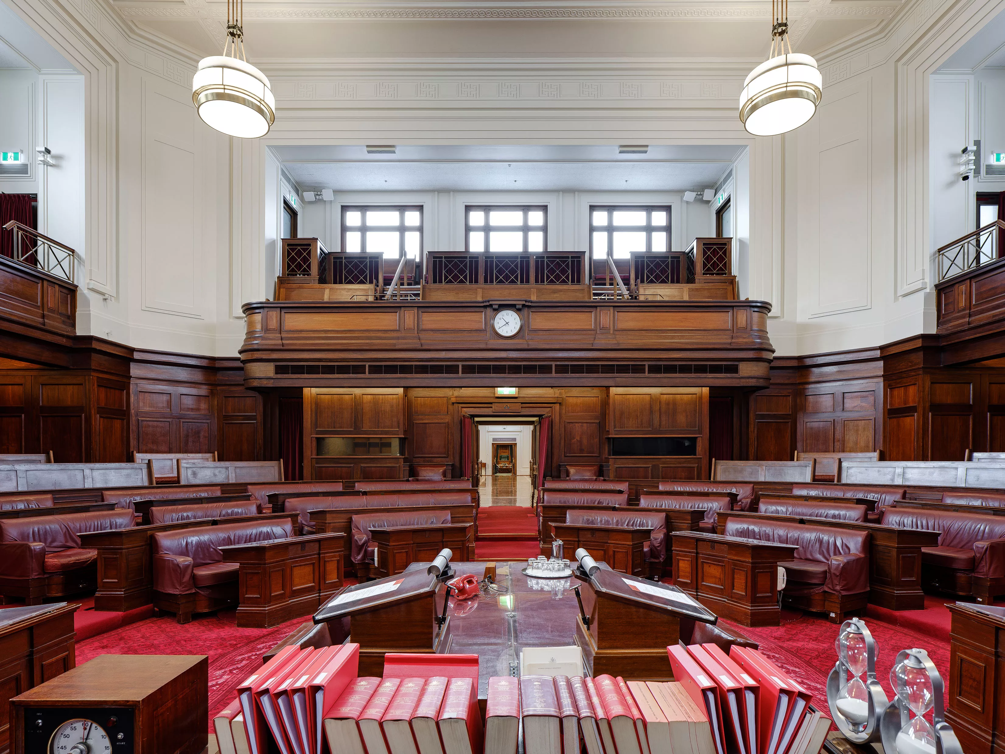 A view out to the Senate chamber. In the foreground are red leather bound books on a long table. Red carpet and red leather chairs in a horseshoe shape with timber panels and art deco lights. 