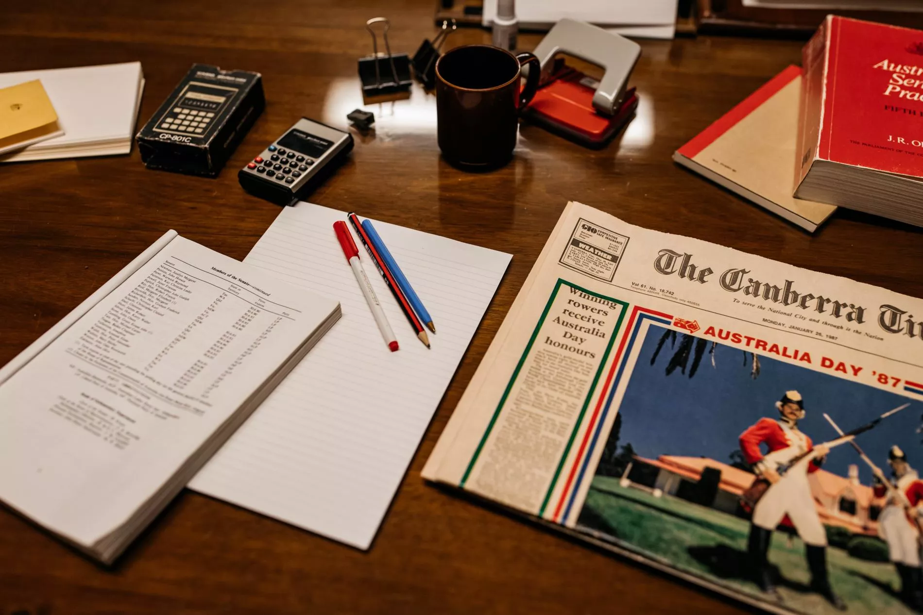 A view of the top of a desk with a Canberra paper from 1987, pens, notepads and an Australian Pubic Service book.