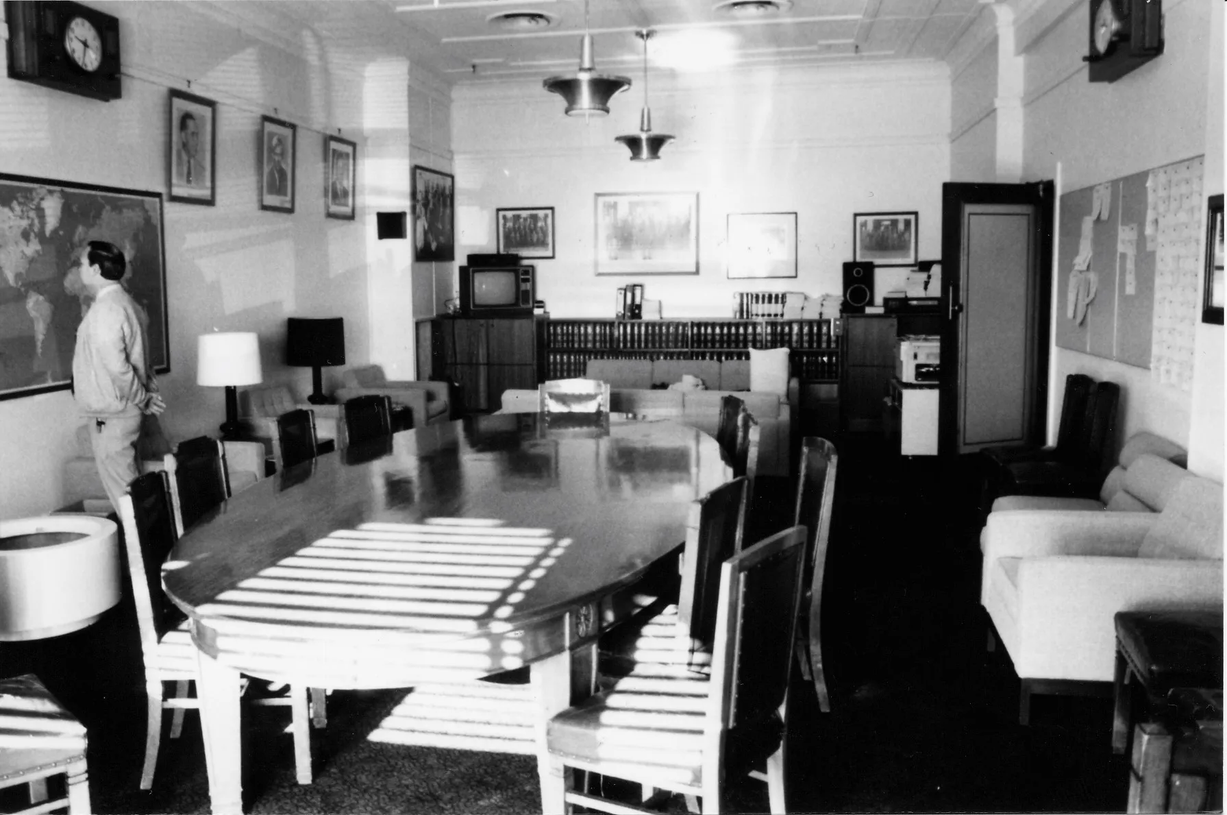 A large oval meeting table and chairs dominate the centre of the room. Easy chairs with side tables and lamps are arrayed along the sides and two sofas are arranged in a seating area towards the back of the room. A sideboard with books, a television and sound system stretches along the back wall. There is a framed map on one wall and a notice board on the opposite. Portraits of leaders and formal and informal photographs of the party crowd the walls A large oval meeting table and chairs dominate the centre of the room. Easy chairs with side tables and lamps are arrayed along the sides and two sofas are arranged in a seating area towards the back of the room. A sideboard with books, a television and sound system stretches along the back wall. There is a framed map on one wall and a notice board on the opposite. Portraits of leaders and formal and informal photographs of the party crowd the walls