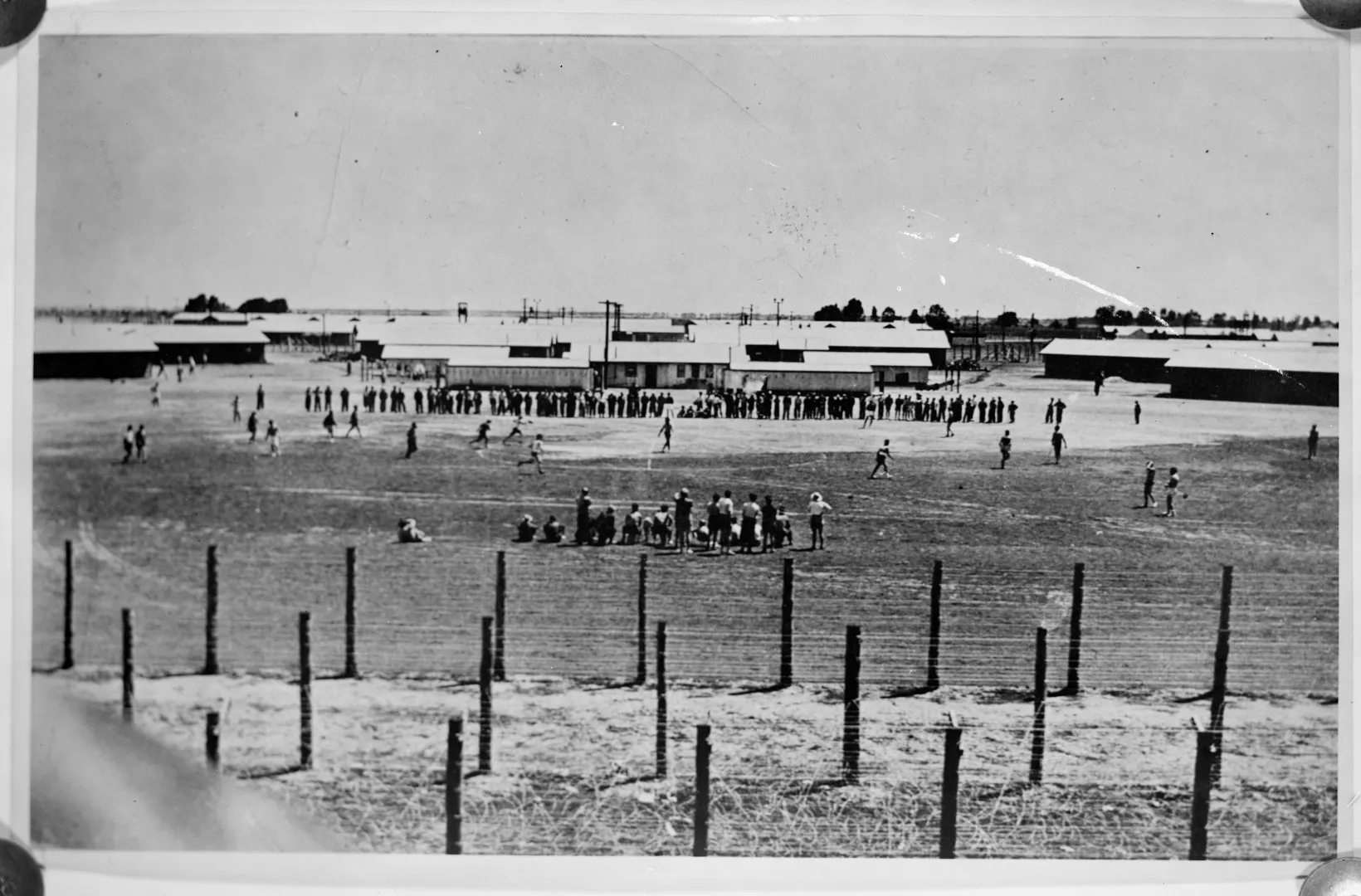 Black and white photograph of Prisoners playing soccer in an internment camp, with military buildings behind them and razor wire in the foreground.  