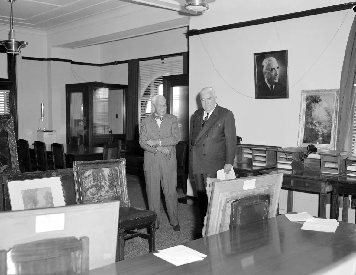 This black and white photograph shows two suited men standing side by side in the Government Party Room in 1962. On the left is Sydney businessman and philanthropist SH Ervin with a jaunty bow tie, on the right a more conservatively dressed Prime Minister Bob Menzies. They are surrounded by Australian paintings which are leaning against chairs, tables and walls. The two men are viewing the paintings that Ervin was donating to the National Collection. In the background are meeting tables and telephone booths This black and white photograph shows two suited men standing side by side in the Government Party Room in 1962. On the left is Sydney businessman and philanthropist SH Ervin with a jaunty bow tie, on the right a more conservatively dressed Prime Minister Bob Menzies. They are surrounded by Australian paintings which are leaning against chairs, tables and walls. The two men are viewing the paintings that Ervin was donating to the National Collection. In the background are meeting tables and telephone booths