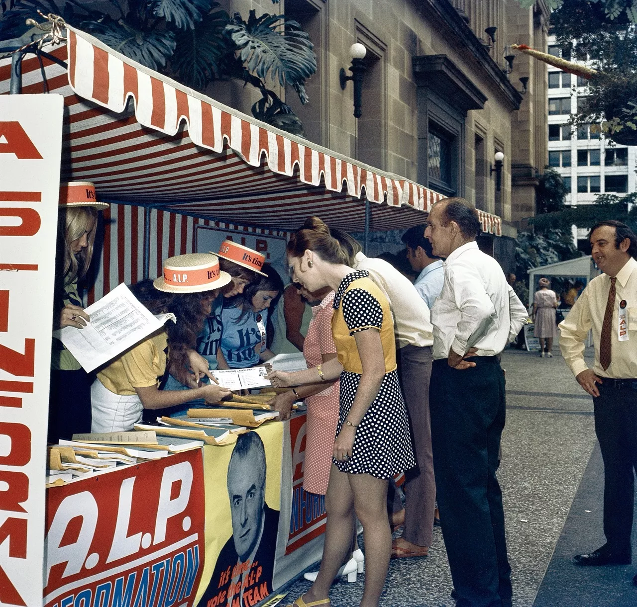 Campaign workers wearing orange bands around their hats at a booth covered in Labor bunting and poster, speaking to voters and showing them leaflets in a city street.     