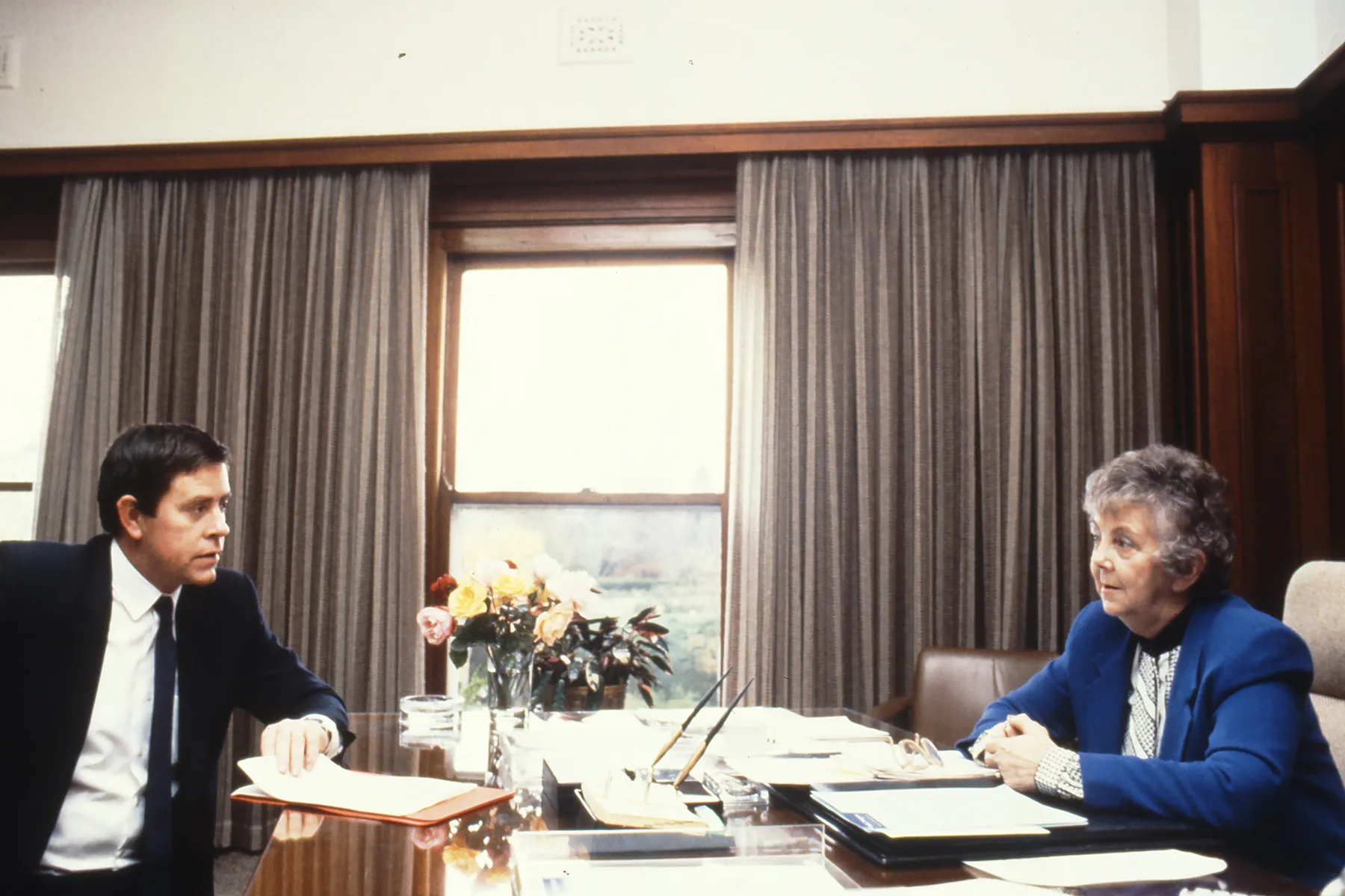 This professional colour photograph shows Principal Private Secretary John Porter and Speaker Joan Child sitting across from each other at the Speaker’s desk. John is wearing a dark suit, white shirt and dark tie and Joan is in a white blouse and vibrant blue jacket. They are discussing the daily program for the House of Representatives Chamber. There is a vase of roses, stationery and paperwork on the desk. In the background the curtains have been drawn to reveal a large east-facing window.