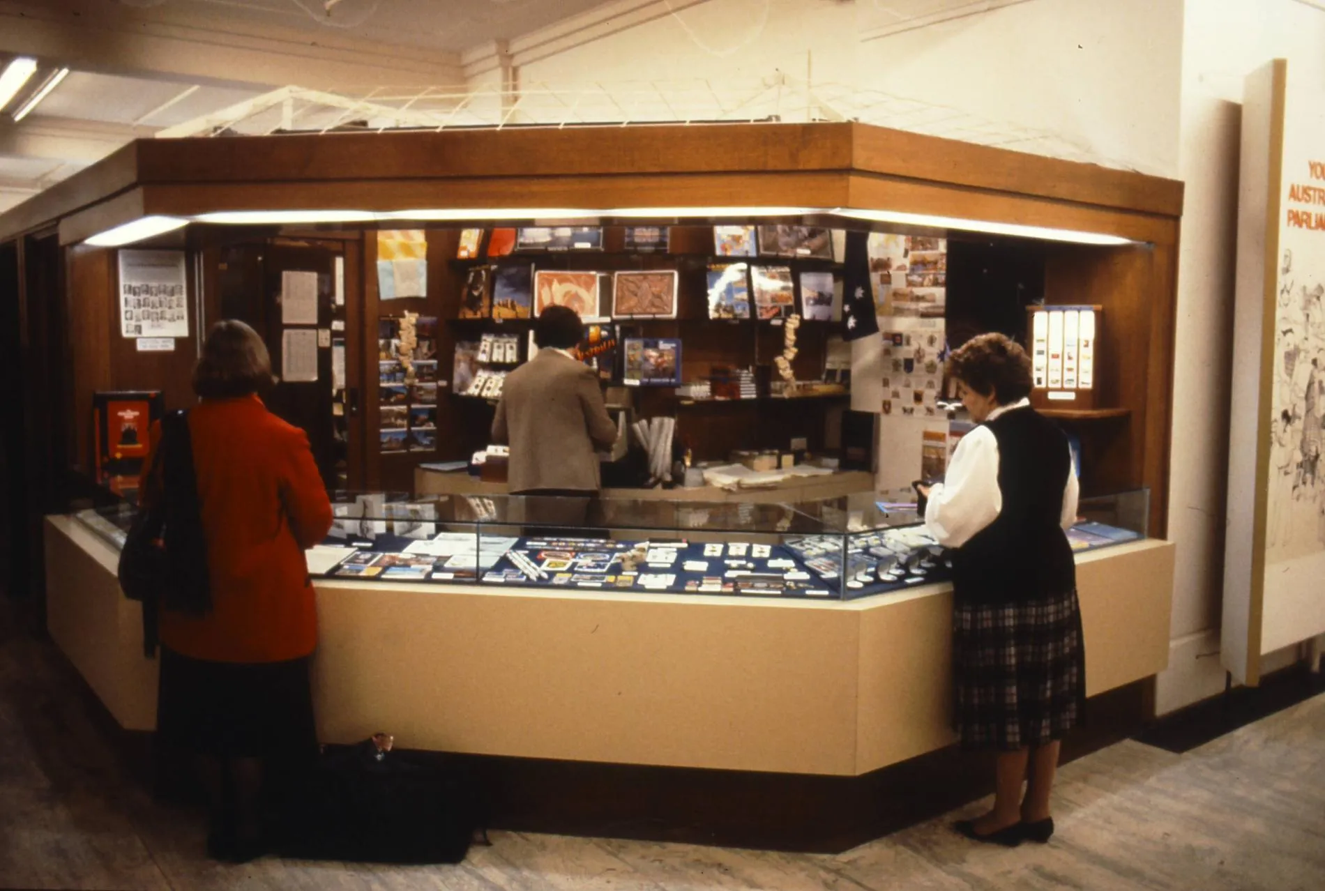 Two women stand at the glass display counter of the Old Parliament House gift shop. A man behind the counter is on a till.