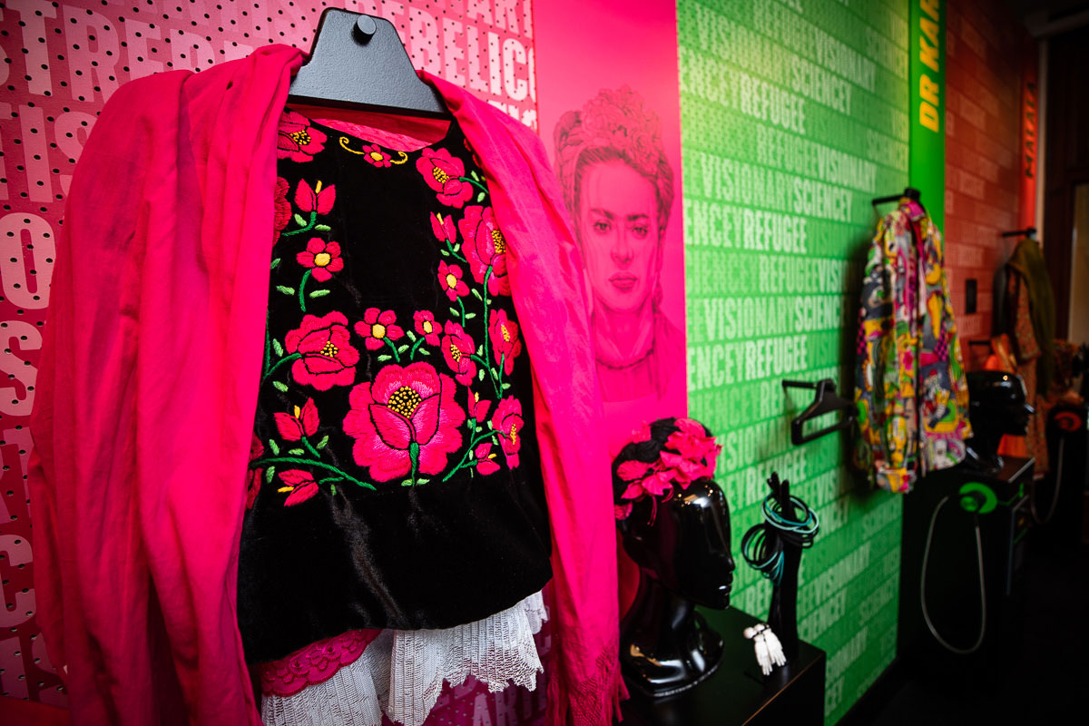 A bright pink coat over a black top with embroidered pink flowers hangs on a pink wall with an image of Frida Kahlo next to it in a room with more costumes to dress up in. A bright pink coat over a black top with embroidered pink flowers hangs on a pink wall with an image of Frida Kahlo next to it in a room with more costumes to dress up in.