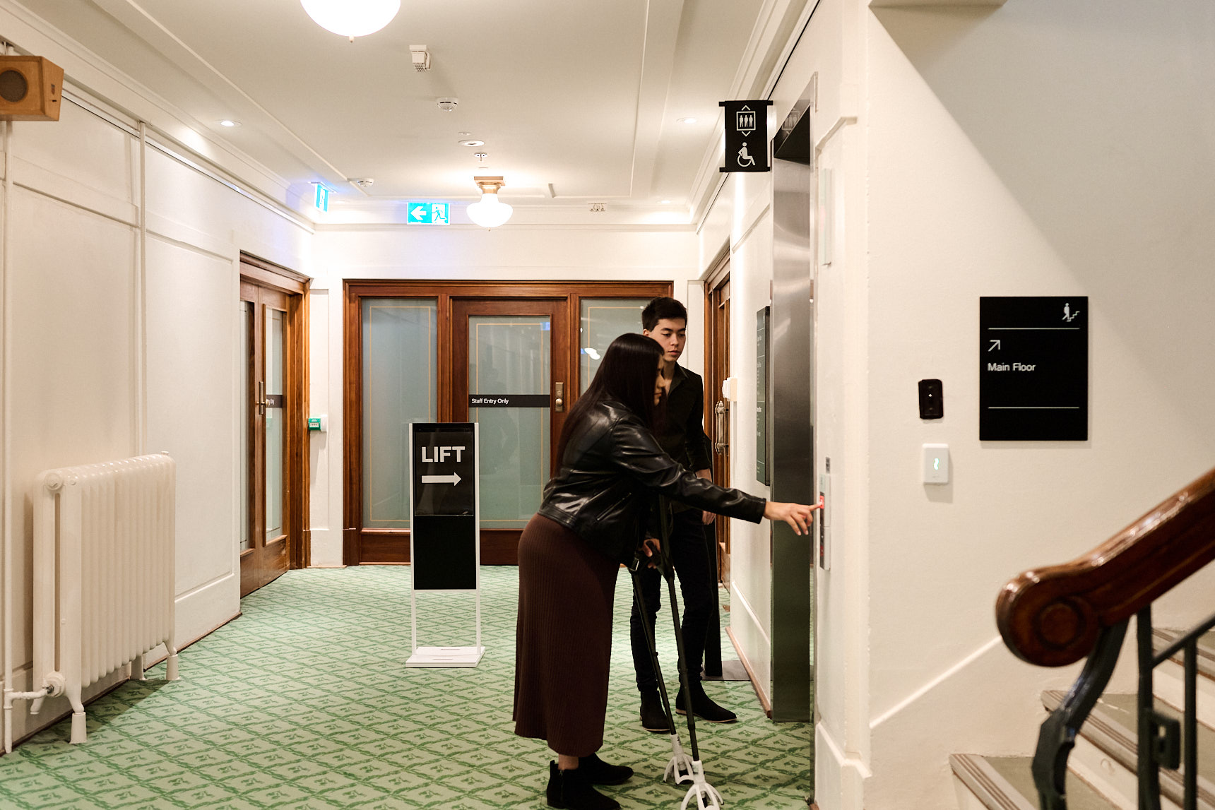 A woman on crutches and a man stand outside a lift on green carpet in Old Parliament House. A woman on crutches and a man stand outside a lift on green carpet in Old Parliament House.