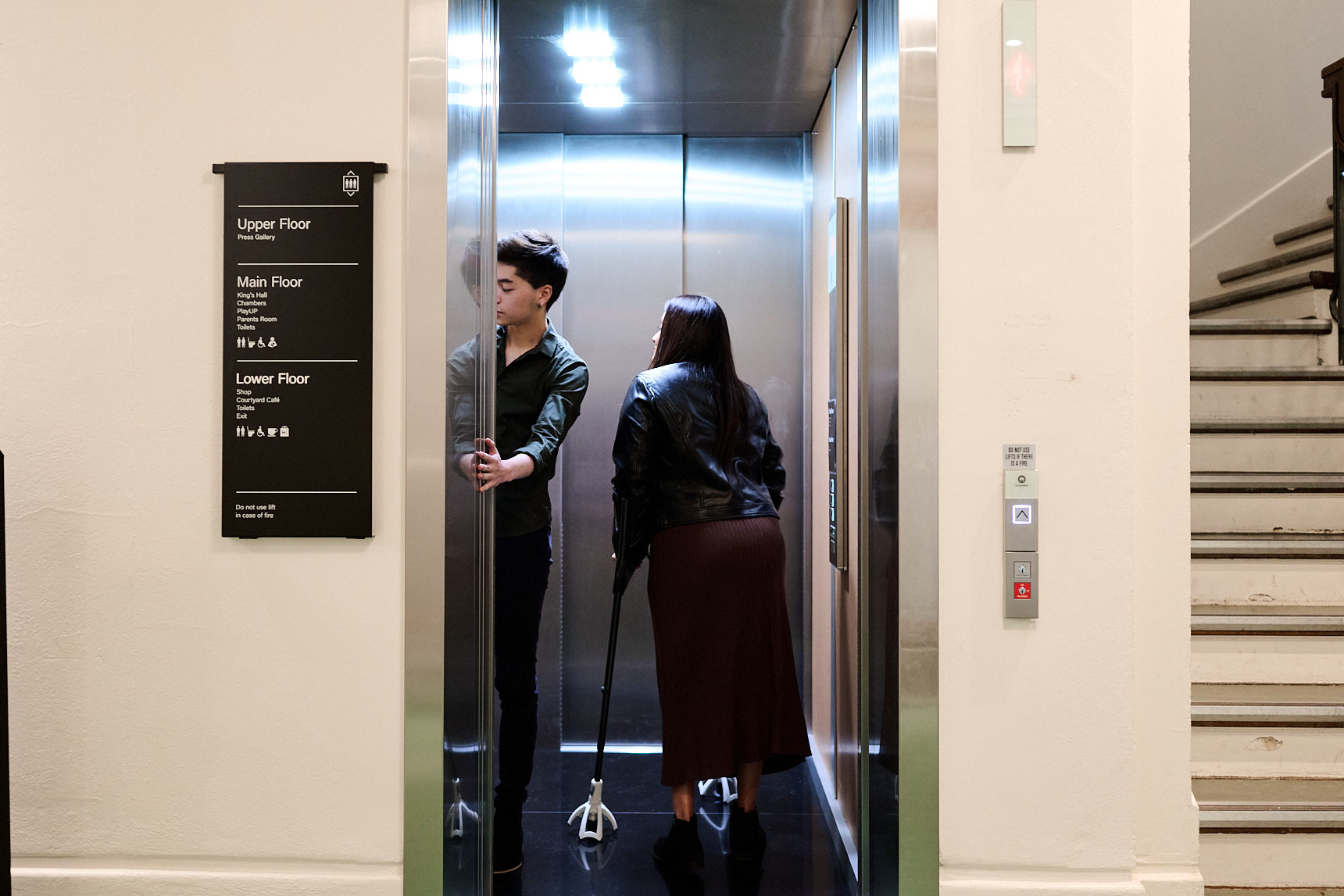 A man holds the door for a woman on crutches inside a lift at Old Parliament House, there is a sign outside the lift for the different floors of the building. A man holds the door for a woman on crutches inside a lift at Old Parliament House, there is a sign outside the lift for the different floors of the building.