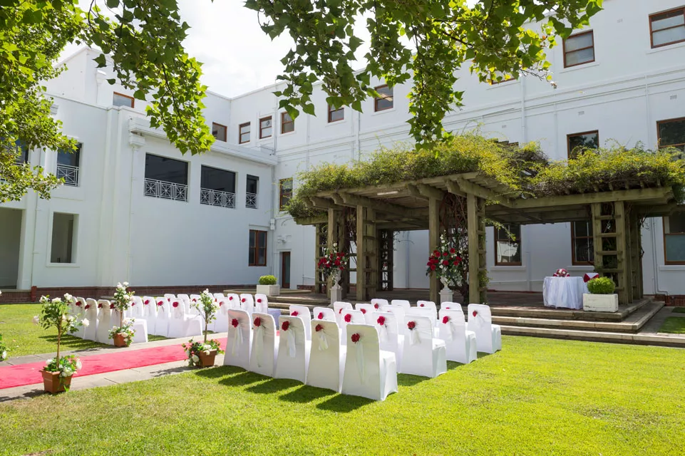 White chairs set up in rows on a lawn facing a wooden pergola covered in vines, ready for a wedding. White chairs set up in rows on a lawn facing a wooden pergola covered in vines, ready for a wedding.