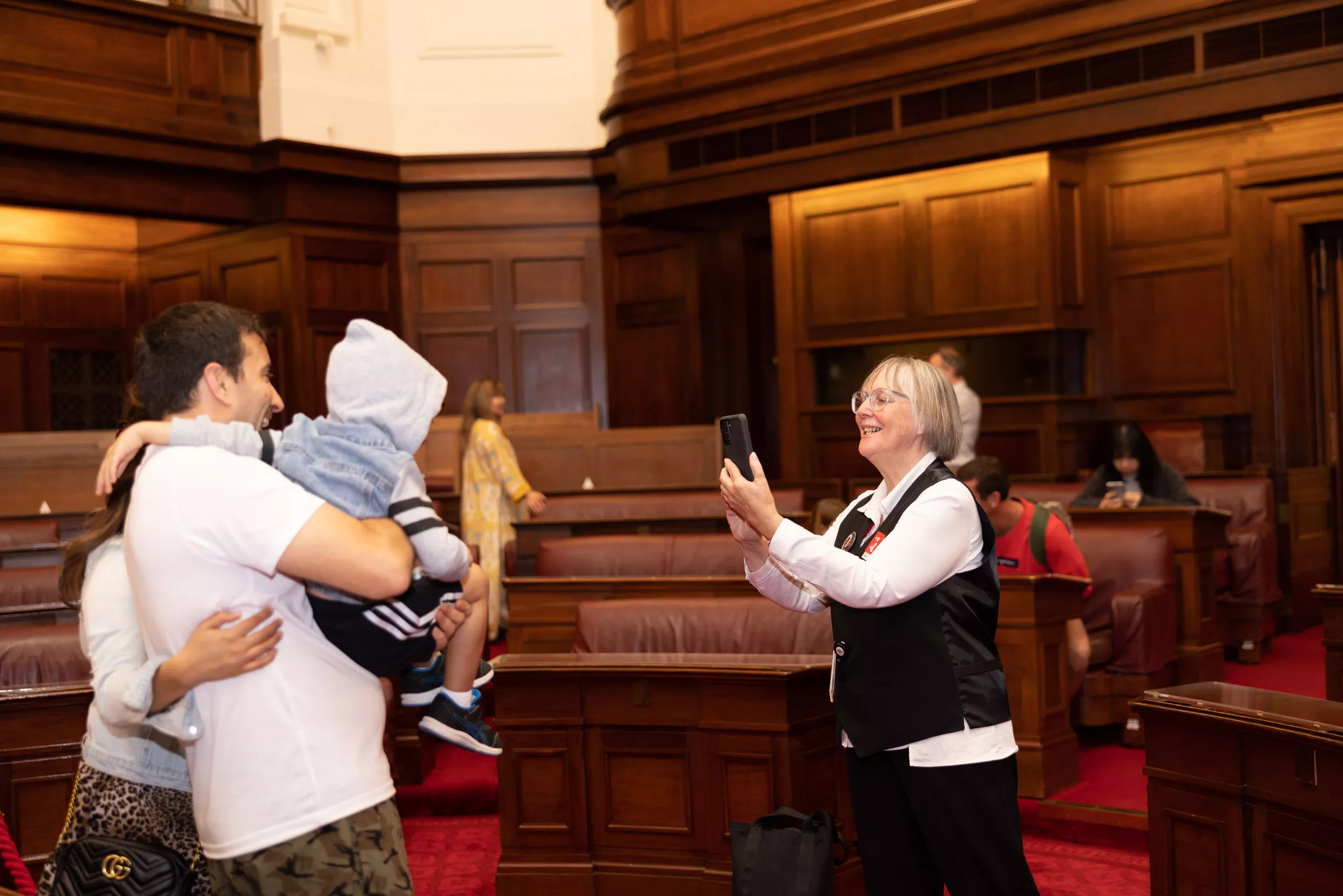 A volunteer takes a photo with a phone of a couple holding a young boy in the Senate Chamber. A volunteer takes a photo with a phone of a couple holding a young boy in the Senate Chamber.