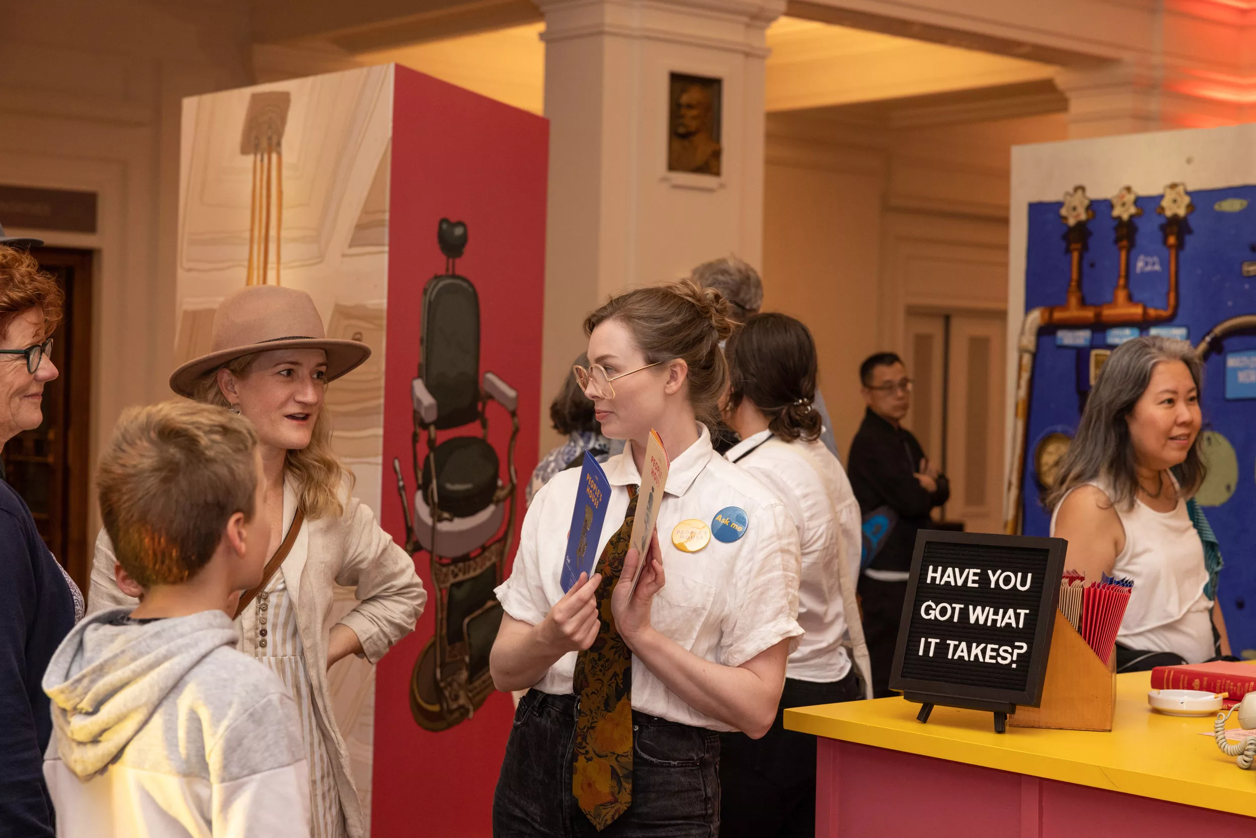 A woman with a white shirt and tie stands in front of a colourful display talking to a woman and young boy and showing them pamphlets. A woman with a white shirt and tie stands in front of a colourful display talking to a woman and young boy and showing them pamphlets.