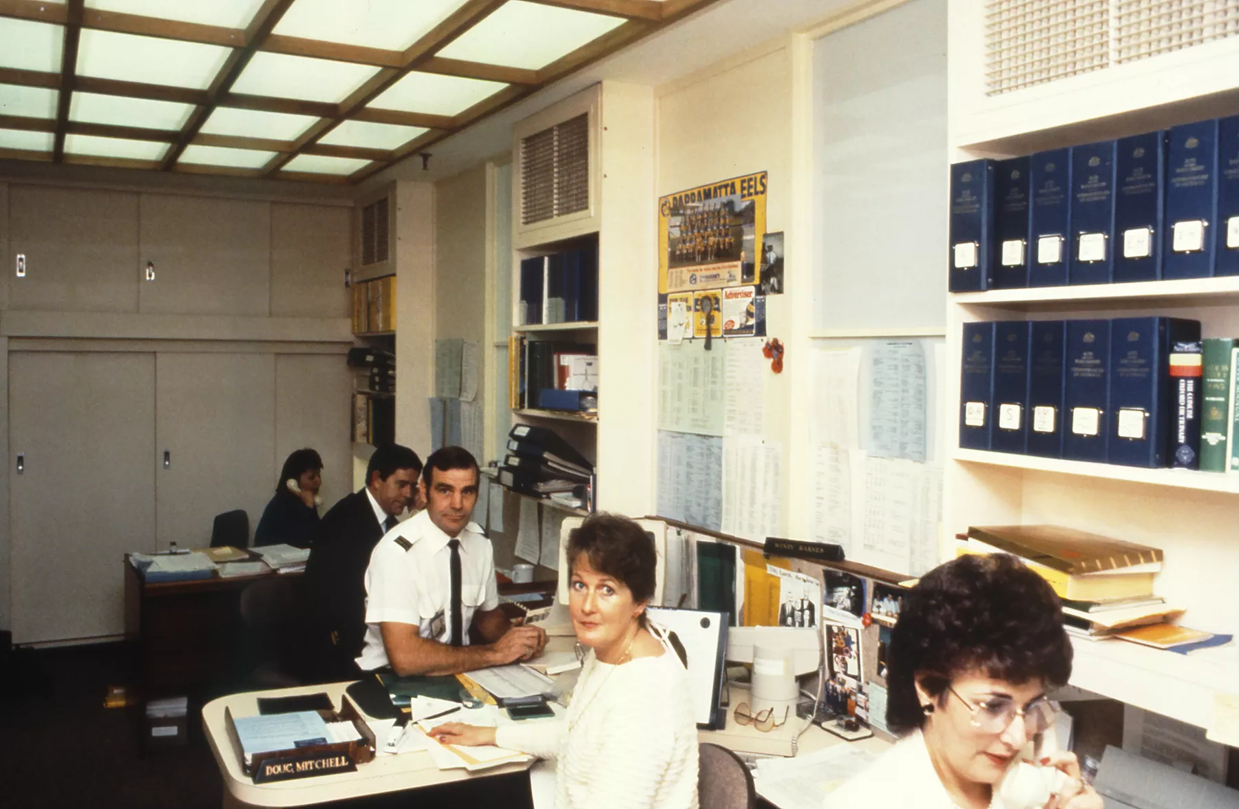 This professional colour photograph shows a busy and crowded open plan office in the Speaker’s Suite in 1988 – just a few months before parliament moved to Australian Parliament House. All five desks are occupied with Private Secretary Koula Alexiadis, Principal Private Secretary John Porter, Attendant Doug Mitchell, Assistant Private Secretary Wendy Barnes and Bookings Officer Marie Donnelly. Koula, John and Marie are all on the telephone while Doug and Wendy smile at the camera. This professional colour photograph shows a busy and crowded open plan office in the Speaker’s Suite in 1988 – just a few months before parliament moved to Australian Parliament House. All five desks are occupied with Private Secretary Koula Alexiadis, Principal Private Secretary John Porter, Attendant Doug Mitchell, Assistant Private Secretary Wendy Barnes and Bookings Officer Marie Donnelly. Koula, John and Marie are all on the telephone while Doug and Wendy smile at the camera.