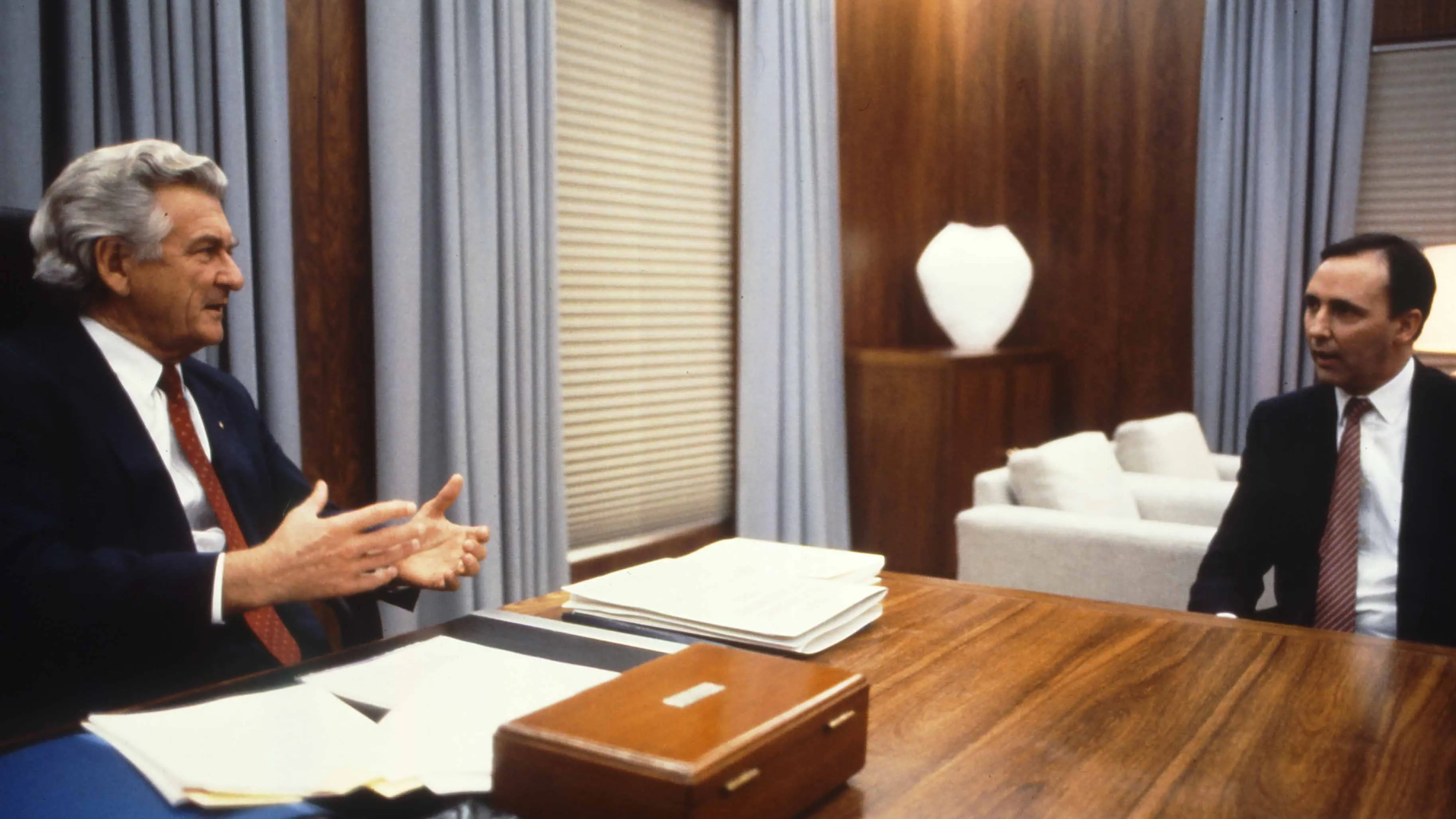 This colour photograph captures a conversation between Prime Minister Bob Hawke seated on the left and Treasurer Paul Keating seated on the right. The photo shows the corner of the desk with Hawke sitting behind the desk while Keating sits to the side with both men facing each other. Both are dressed in black suits with white shirts and red ties contrasting the pale blue curtains in the background. This colour photograph captures a conversation between Prime Minister Bob Hawke seated on the left and Treasurer Paul Keating seated on the right. The photo shows the corner of the desk with Hawke sitting behind the desk while Keating sits to the side with both men facing each other. Both are dressed in black suits with white shirts and red ties contrasting the pale blue curtains in the background.