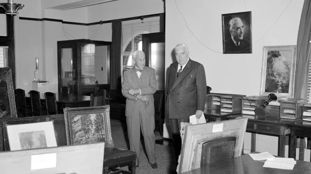 This black and white photograph shows two suited men standing side by side in the Government Party Room in 1962. On the left is Sydney businessman and philanthropist SH Ervin with a jaunty bow tie, on the right a more conservatively dressed Prime Minister Bob Menzies. They are surrounded by Australian paintings which are leaning against chairs, tables and walls. The two men are viewing the paintings that Ervin was donating to the National Collection. In the background are meeting tables and telephone booths This black and white photograph shows two suited men standing side by side in the Government Party Room in 1962. On the left is Sydney businessman and philanthropist SH Ervin with a jaunty bow tie, on the right a more conservatively dressed Prime Minister Bob Menzies. They are surrounded by Australian paintings which are leaning against chairs, tables and walls. The two men are viewing the paintings that Ervin was donating to the National Collection. In the background are meeting tables and telephone booths