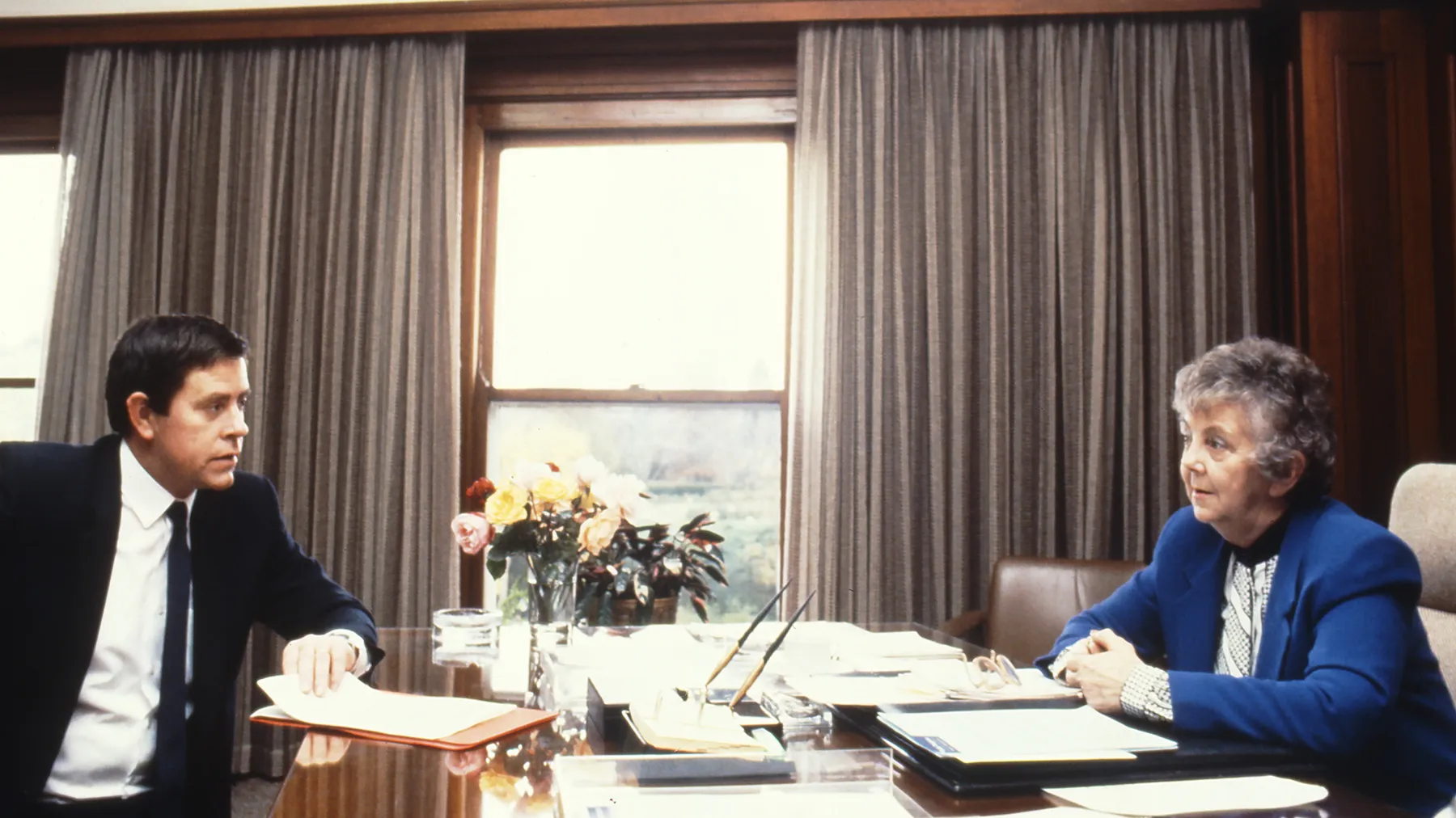This professional colour photograph shows Principal Private Secretary John Porter and Speaker Joan Child sitting across from each other at the Speaker’s desk. John is wearing a dark suit, white shirt and dark tie and Joan is in a white blouse and vibrant blue jacket. They are discussing the daily program for the House of Representatives Chamber. There is a vase of roses, stationery and paperwork on the desk. In the background the curtains have been drawn to reveal a large east-facing window. This professional colour photograph shows Principal Private Secretary John Porter and Speaker Joan Child sitting across from each other at the Speaker’s desk. John is wearing a dark suit, white shirt and dark tie and Joan is in a white blouse and vibrant blue jacket. They are discussing the daily program for the House of Representatives Chamber. There is a vase of roses, stationery and paperwork on the desk. In the background the curtains have been drawn to reveal a large east-facing window.