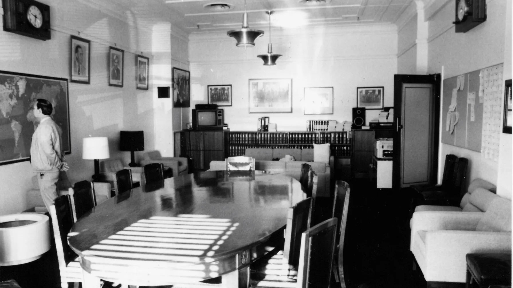 A large oval meeting table and chairs dominate the centre of the room. Easy chairs with side tables and lamps are arrayed along the sides and two sofas are arranged in a seating area towards the back of the room. A sideboard with books, a television and sound system stretches along the back wall. There is a framed map on one wall and a notice board on the opposite. Portraits of leaders and formal and informal photographs of the party crowd the walls A large oval meeting table and chairs dominate the centre of the room. Easy chairs with side tables and lamps are arrayed along the sides and two sofas are arranged in a seating area towards the back of the room. A sideboard with books, a television and sound system stretches along the back wall. There is a framed map on one wall and a notice board on the opposite. Portraits of leaders and formal and informal photographs of the party crowd the walls