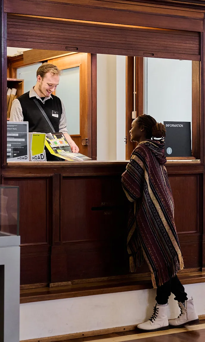 A Museum Experience Officer wearing a black vest and name tag standing inside an information booth at MoAD at Old Parliament House assists a woman with a brochure about the Museum. A Museum Experience Officer wearing a black vest and name tag standing inside an information booth at MoAD at Old Parliament House assists a woman with a brochure about the Museum.