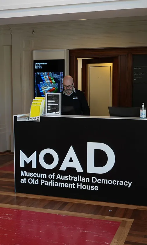 A museum staff member wearing a black sweater and name badge stands behind the main entrance reception desk at MoAD, the Museum of Australian Democracy at Old Parliament House. A museum staff member wearing a black sweater and name badge stands behind the main entrance reception desk at MoAD, the Museum of Australian Democracy at Old Parliament House.