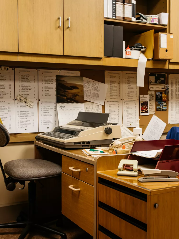 A 1980s looking office with brown desks, typewriters and printed pages stuck to a corkboard.
