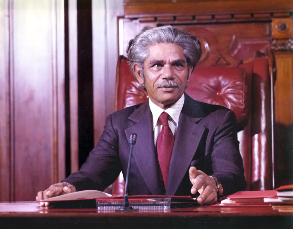 Neville Bonner seated in the Senate chamber, wearing a suit and red tie. Neville Bonner seated in the Senate chamber, wearing a suit and red tie.