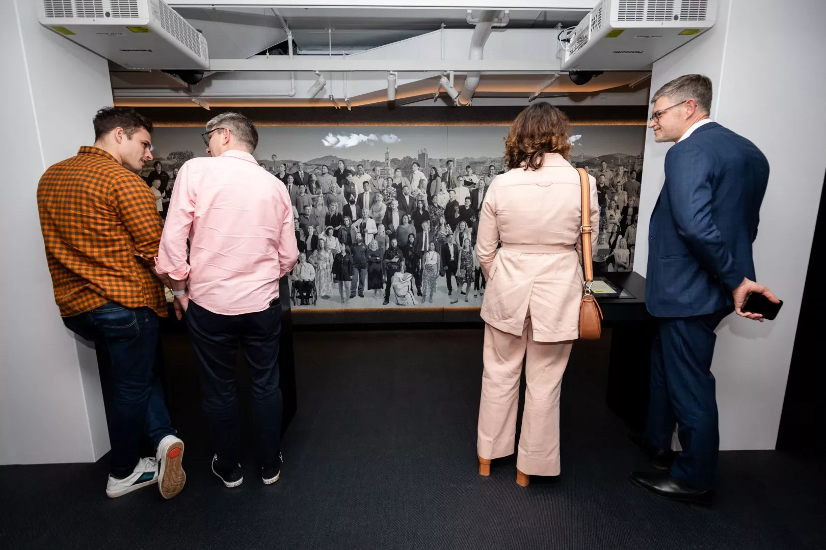 A man leans against a pillar talking to another man. In front of them is a large black and white photo featuring lots of different people. A man leans against a pillar talking to another man. In front of them is a large black and white photo featuring lots of different people.