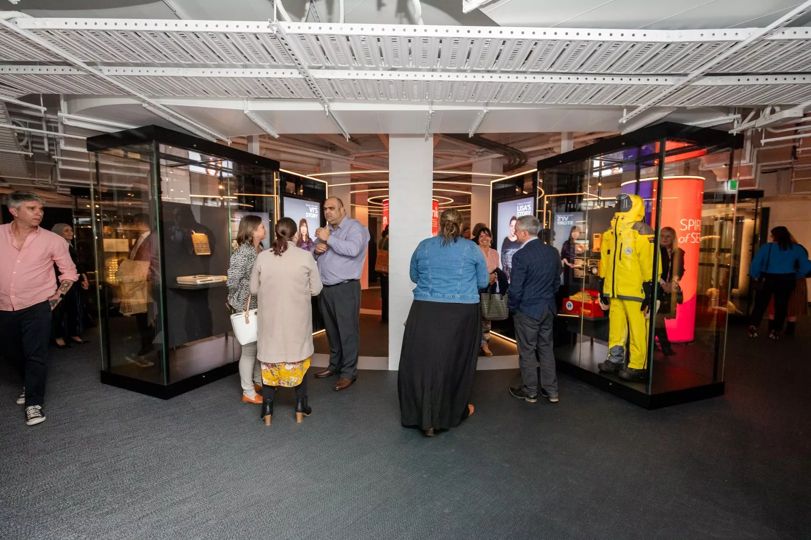 People mill around talking to each other and looking into glass displays in a museum exhibition. One features a bright yellow suit. People mill around talking to each other and looking into glass displays in a museum exhibition. One features a bright yellow suit.